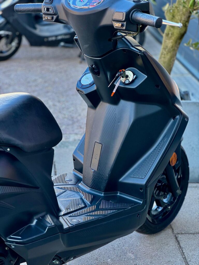 Close-up van een zwarte scooter met sleutel in het contact, gezien bij stuur en voetplaat op straat.