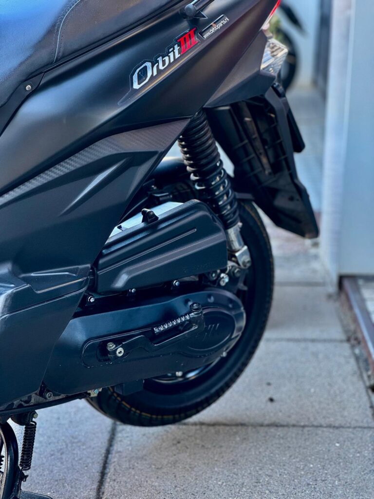Close-up van de achterkant van een zwarte scooter, model Orbit III, met veer en achterwiel op stoeptegels.