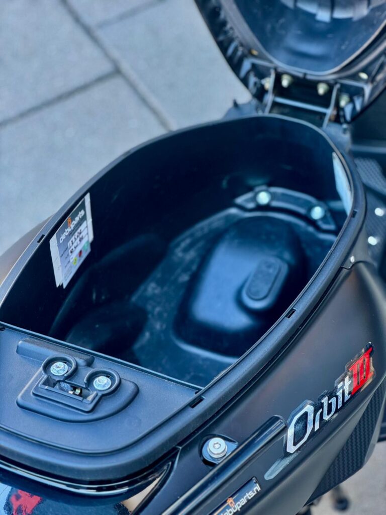 Open opbergruimte onder het zadel van een zwarte scooter, model Orbit III.