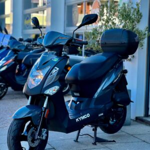 Donkergrijze Kymco-scooter met topkoffer op de standaard, geparkeerd voor een showroom met andere scooters.