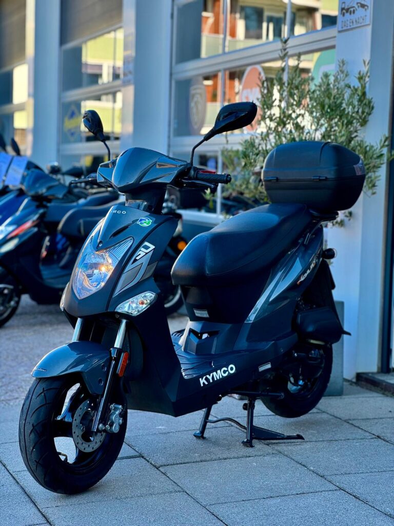 Donkergrijze Kymco-scooter met topkoffer op de standaard, geparkeerd voor een showroom met andere scooters.