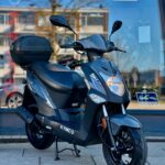 Donkergrijze Kymco-scooter met topkoffer geparkeerd op de stoep voor een winkelraam.