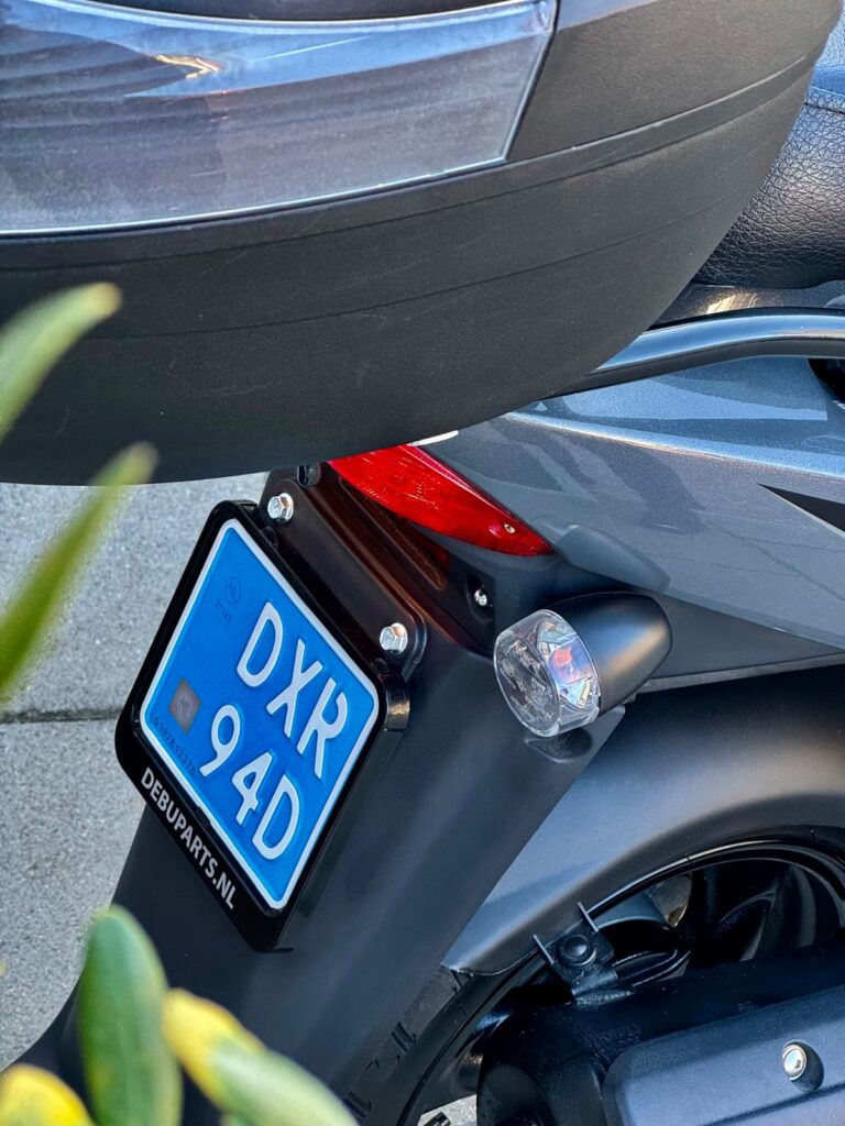 Achterkant van een grijze scooter met blauw kenteken, achterlicht en knipperlicht.