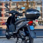Grijze Kymco-scooter met topkoffer, achteraanzicht, geparkeerd op standaard voor een winkelraam.