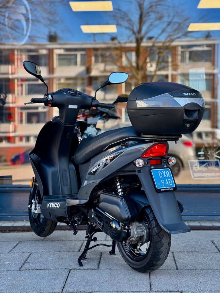 Grijze Kymco-scooter met topkoffer, achteraanzicht, geparkeerd op standaard voor een winkelraam.