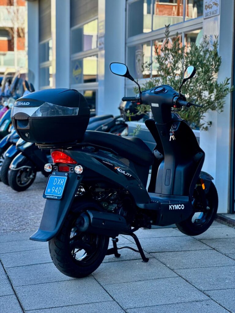 Zwarte Kymco Agility scooter met topkoffer, op middenbok geparkeerd bij showroom.