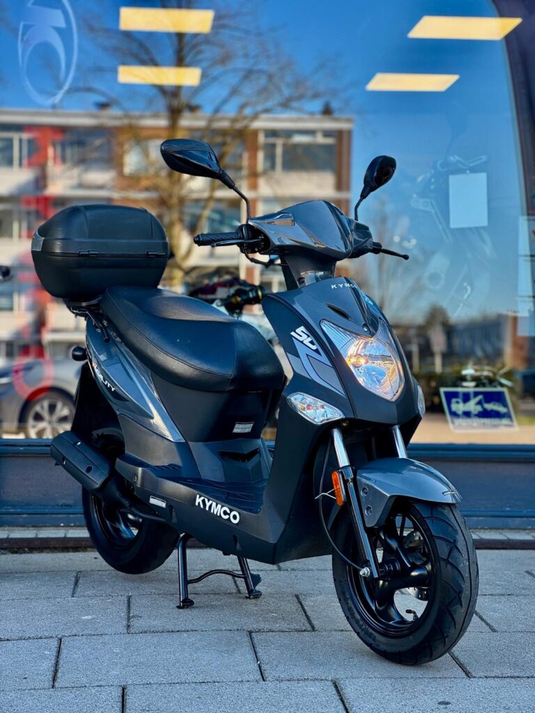 Donkergrijze Kymco-scooter met topkoffer geparkeerd op de stoep voor een winkelraam.