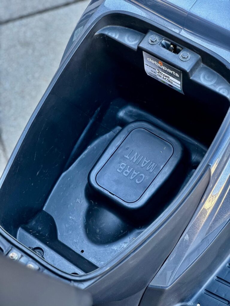 Open opbergruimte onder het zadel van een scooter met CARB. MAINT-klep en label.