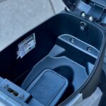 Open zadel van een scooter met opbergruimte, zichtbaar CARB. MAINT.-klep en sticker.