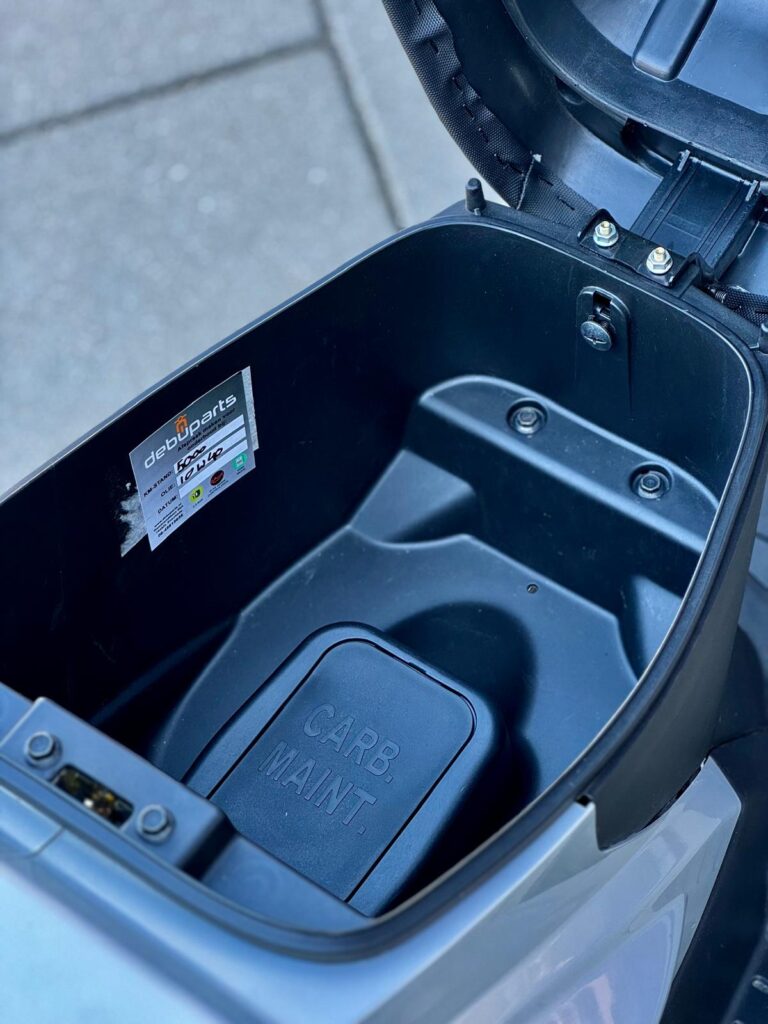 Open zadel van een scooter met opbergruimte, zichtbaar CARB. MAINT.-klep en sticker.