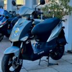 Grijze Kymco-scooter op de middenbok voor een showroom, met voorlamp en schijfrem.