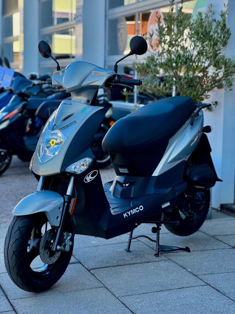 Grijze Kymco-scooter op de middenbok voor een showroom, met voorlamp en schijfrem.