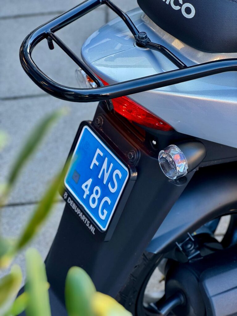 Achterkant van een grijze scooter met blauw kenteken, bagagedrager en achterlicht.