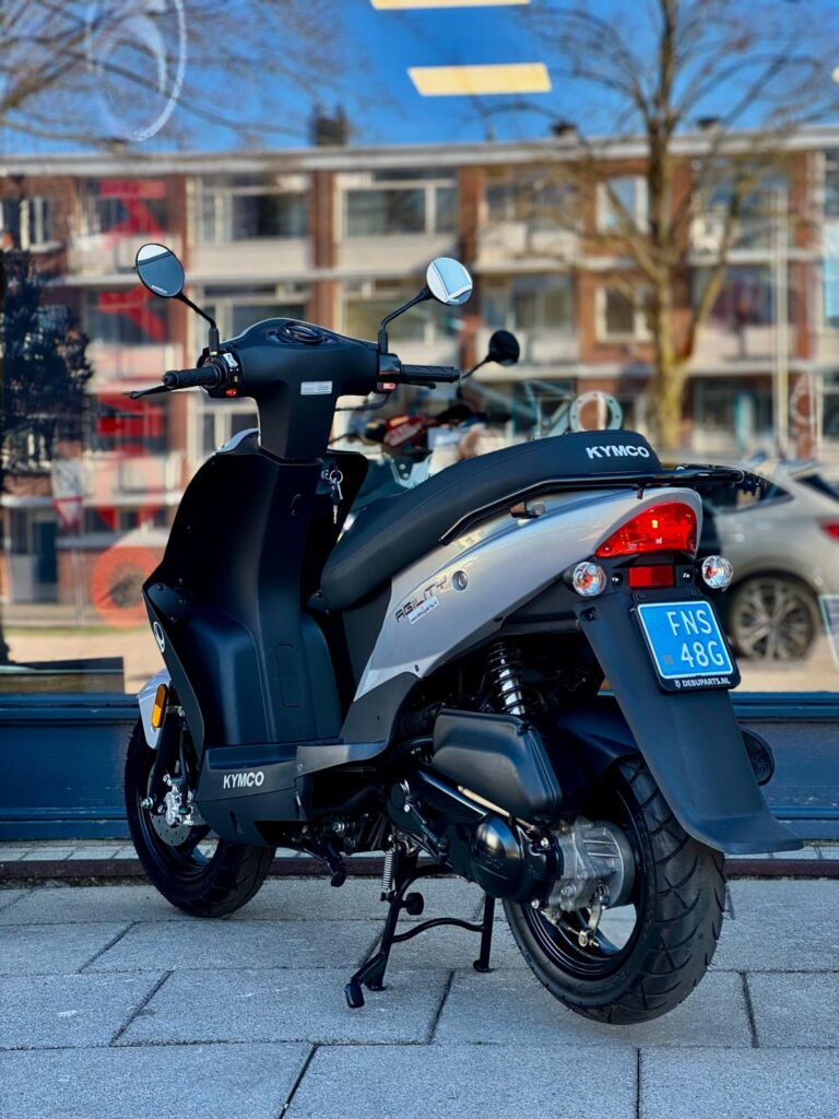 Zwarte en zilveren Kymco Agility scooter op standaard, geparkeerd op stoep voor een etalage.