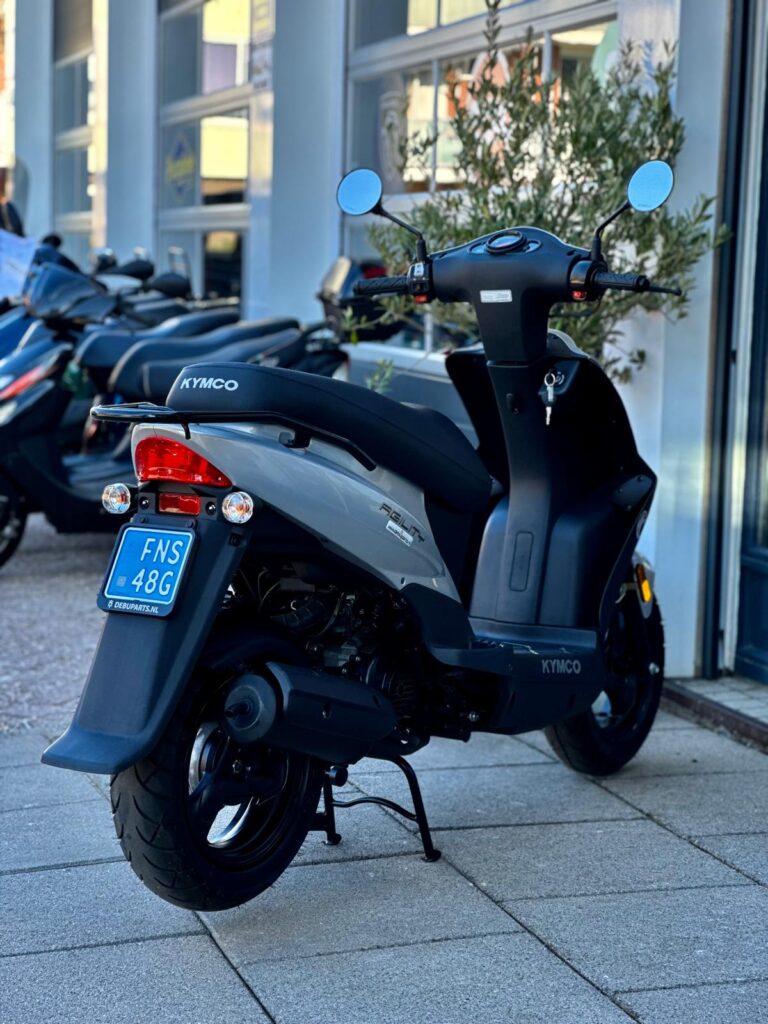 Grijze Kymco-scooter geparkeerd op de stoep voor een winkel, met andere scooters op de achtergrond.