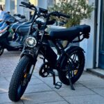 Zwarte fatbike e-bike met dikke banden, geparkeerd op een stoep bij een winkel.