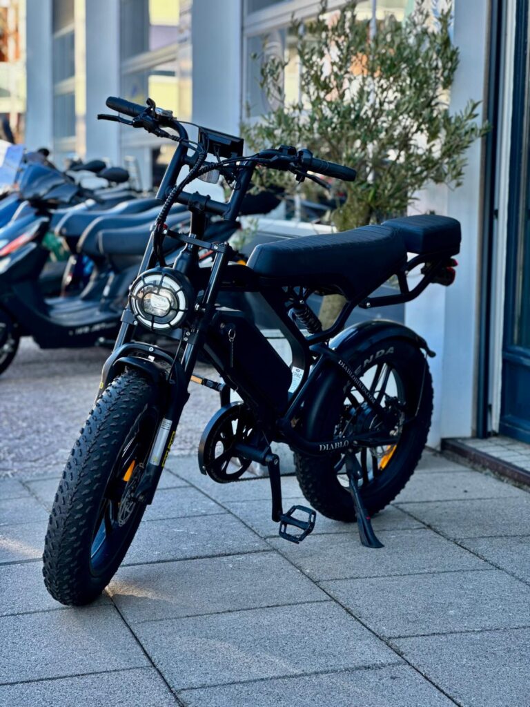 Zwarte fatbike e-bike met dikke banden, geparkeerd op een stoep bij een winkel.