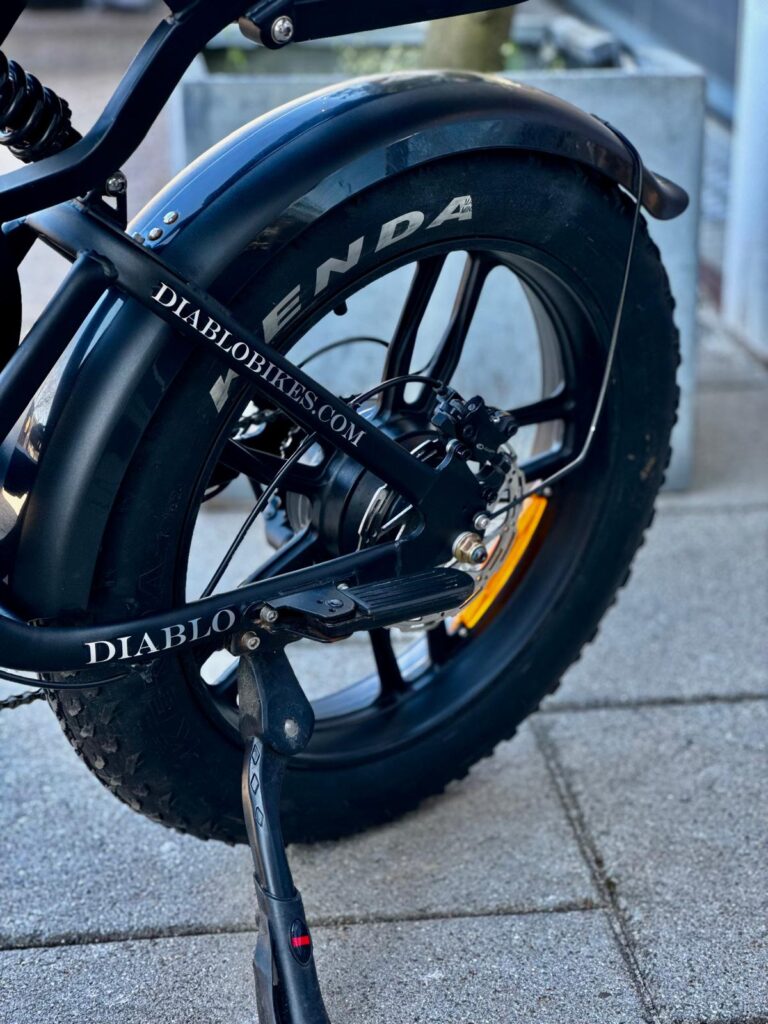 Close-up van het achterwiel van een zwarte fatbike met spatbord, schijfrem en standaard.