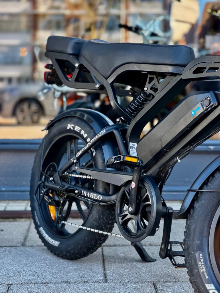 Close-up van een zwarte e-bike met fatbike-banden, dubbele zitting, vering en accupack in het frame.