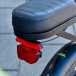 Close-up van een e-bikezadel met rood achterlicht en zwart spatbord, gefotografeerd op straat.