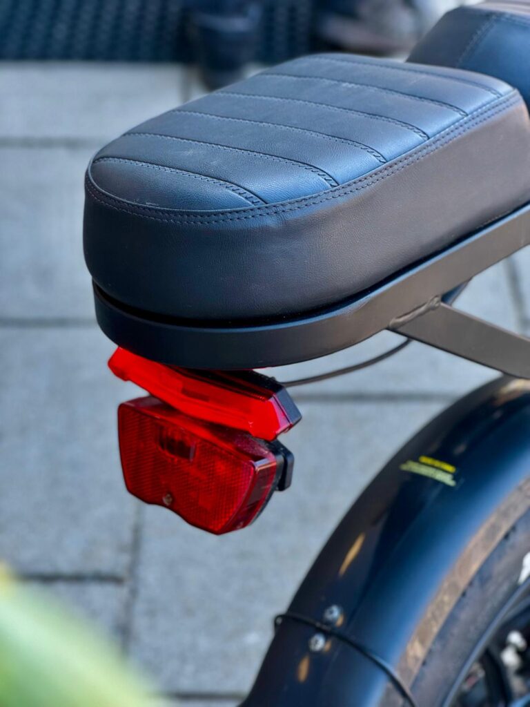 Close-up van een e-bikezadel met rood achterlicht en zwart spatbord, gefotografeerd op straat.