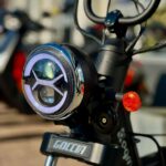 Close-up van koplamp, stuur en oranje richtingaanwijzer van een zwarte Goccia e-scooter.