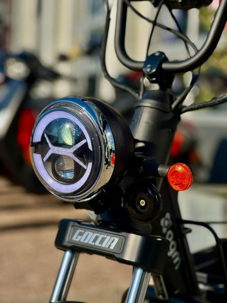 Close-up van koplamp, stuur en oranje richtingaanwijzer van een zwarte Goccia e-scooter.