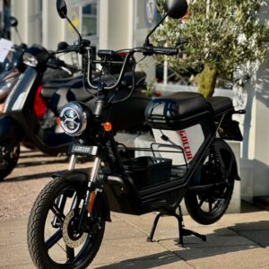 Zwarte elektrische bromfiets met ronde koplamp en mand, geparkeerd voor een scooterzaak.