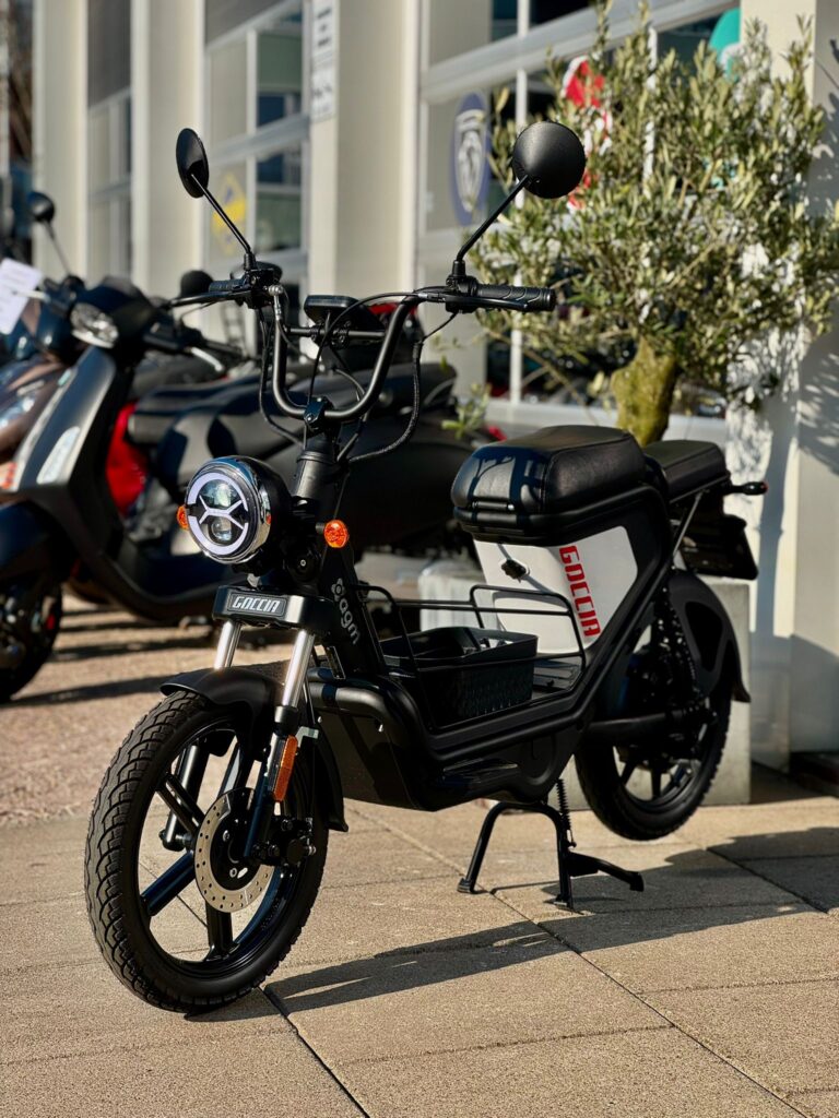 Zwarte elektrische bromfiets met ronde koplamp en mand, geparkeerd voor een scooterzaak.