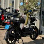 Zwarte elektrische scooter met spiegels op standaard, geparkeerd voor een winkel op zonnige dag.