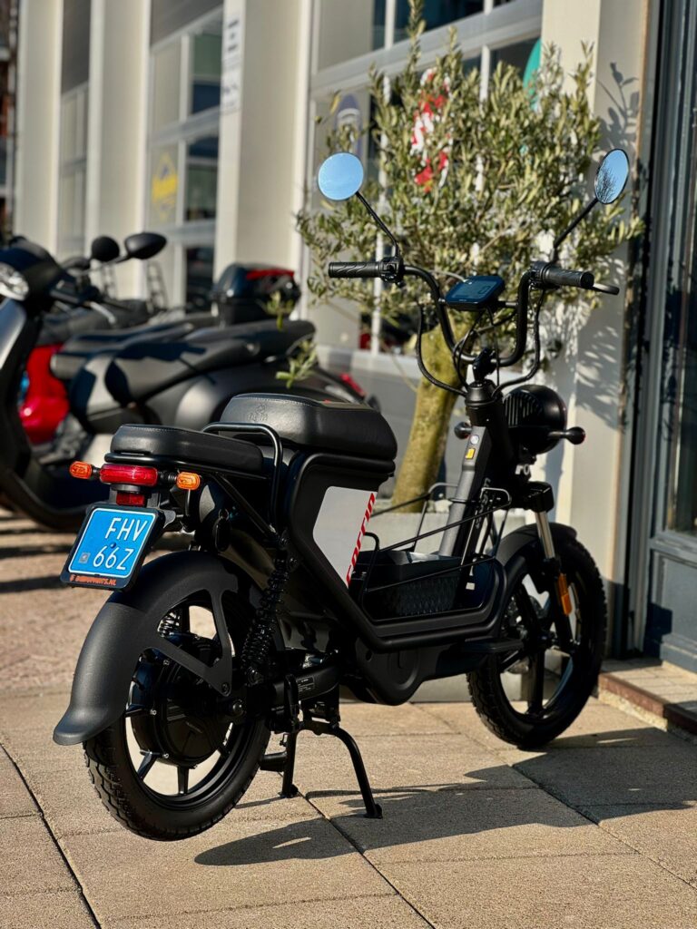 Zwarte elektrische scooter met spiegels op standaard, geparkeerd voor een winkel op zonnige dag.