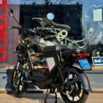 Zwarte elektrische bromfiets met blauw NL-kenteken, geparkeerd voor een winkelruit.