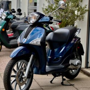 Blauwe scooter op standaard geparkeerd voor een winkel, met andere scooters op de achtergrond.