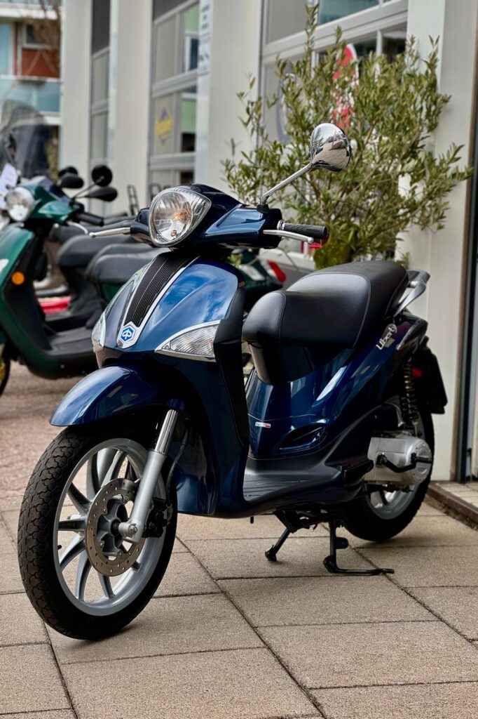 Blauwe scooter op standaard geparkeerd voor een winkel, met andere scooters op de achtergrond.