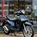 Donkerblauwe Kymco-scooter op standaard voor een winkelruit met weerspiegelde flats.