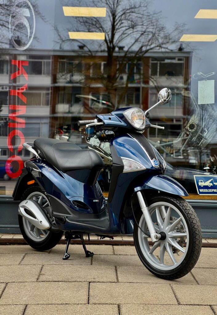 Donkerblauwe Kymco-scooter op standaard voor een winkelruit met weerspiegelde flats.