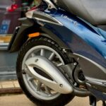 Close-up van het achterwiel, de uitlaat en het zadel van een blauwe Liberty-scooter op de standaard.