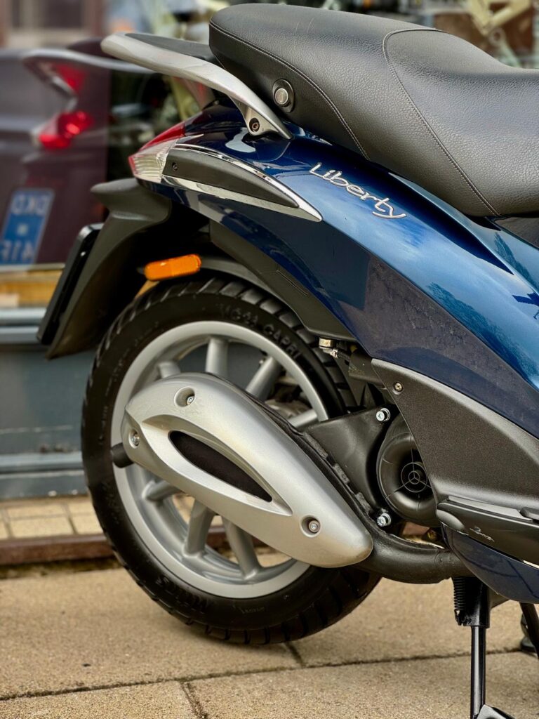 Close-up van het achterwiel, de uitlaat en het zadel van een blauwe Liberty-scooter op de standaard.