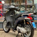 Donkerblauwe scooter op standaard met NL-kenteken, voor een winkelraam geparkeerd.