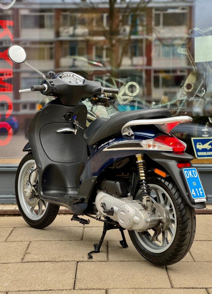 Donkerblauwe scooter op standaard met NL-kenteken, voor een winkelraam geparkeerd.
