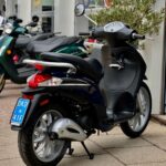 Zwart-blauwe scooter op zijstandaard met blauw kenteken, geparkeerd voor een showroom met scooters.