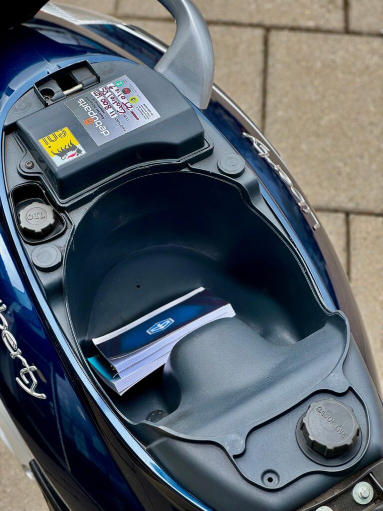 Opbergvak onder het zadel van een scooter met brandstofdop en handleiding.