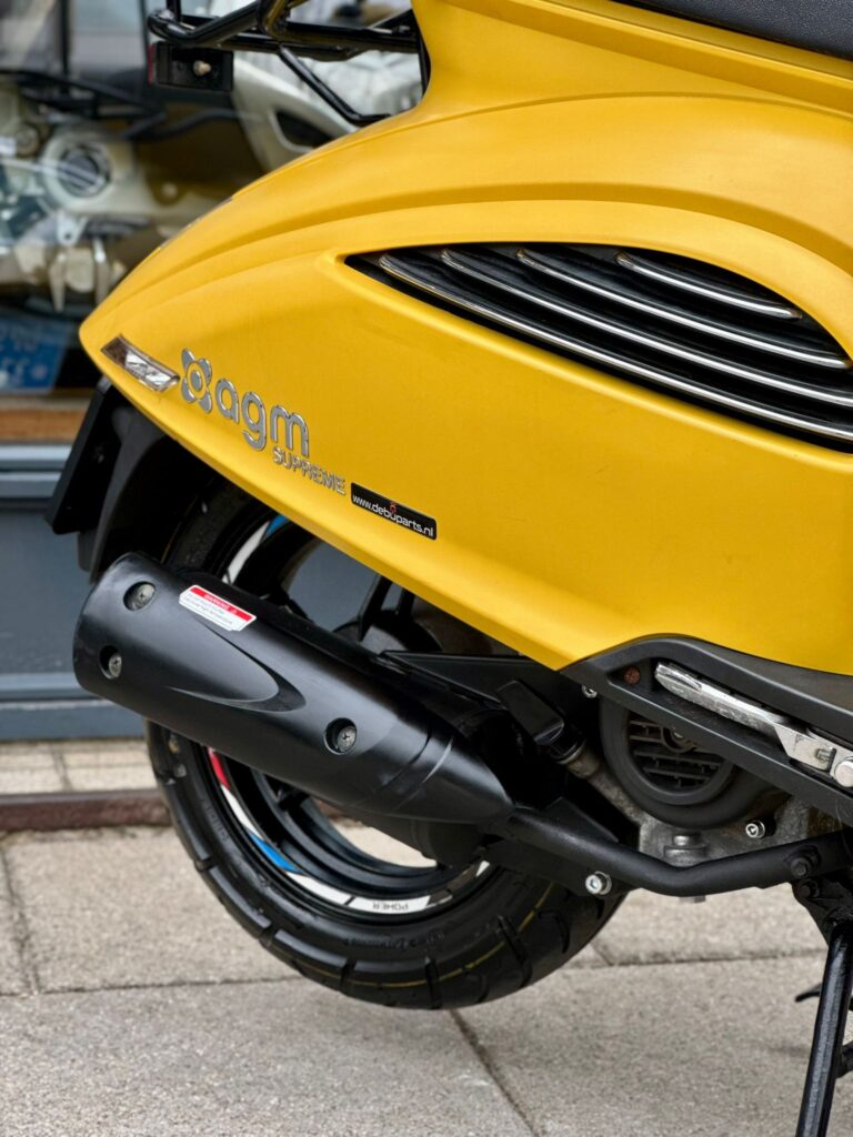 Close-up van de rechterachterzijde van een gele AGM-scooter met uitlaat en achterwiel.