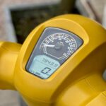 Close-up van het dashboard van een gele scooter met analoge snelheidsmeter en digitaal display.