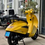 Gele scooter met rugsteun en spiegels geparkeerd op trottoir voor een werkplaats.