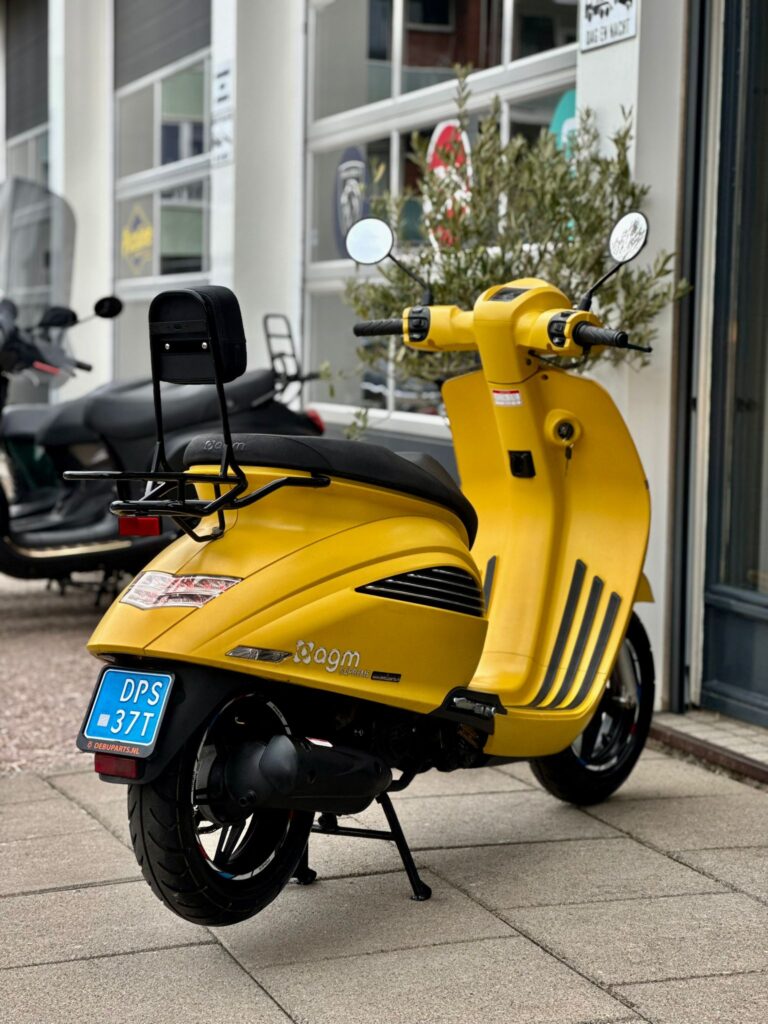 Gele scooter met rugsteun en spiegels geparkeerd op trottoir voor een werkplaats.
