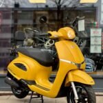 Gele scooter met zwart zadel, op standaard geparkeerd voor een winkelruit