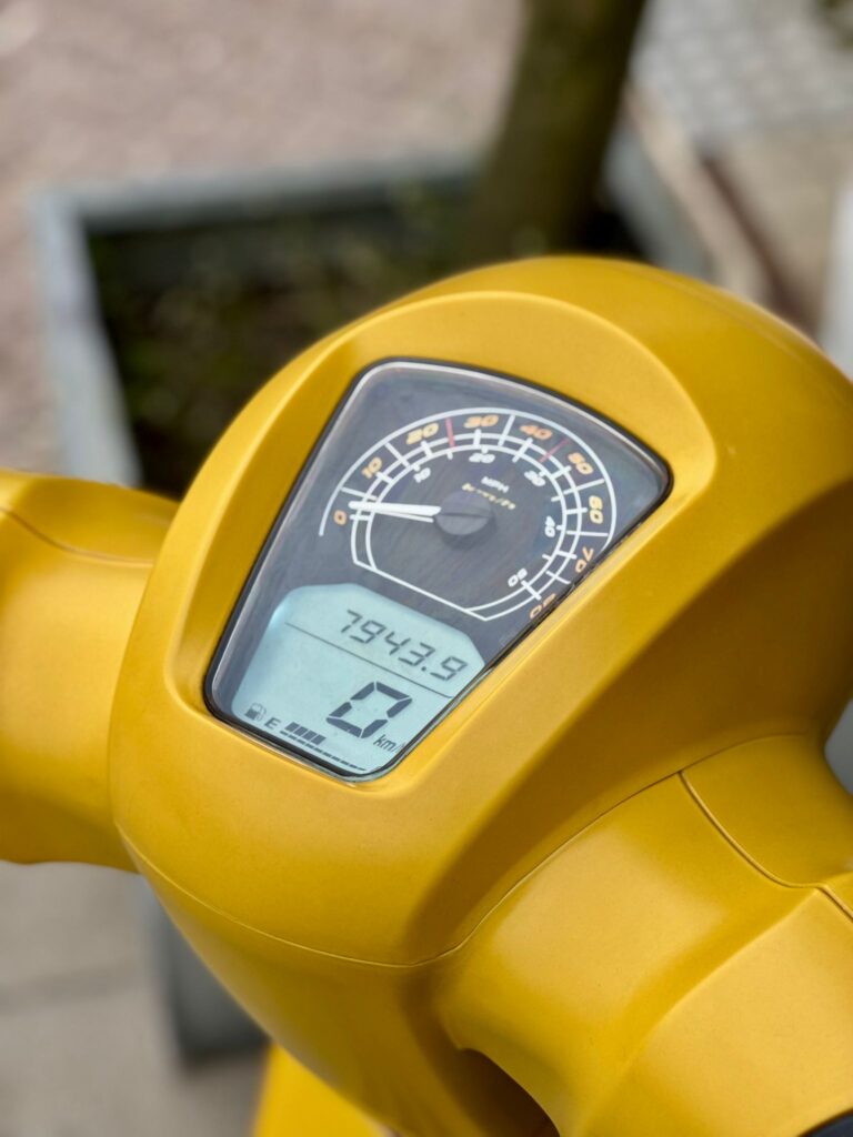 Close-up van het dashboard van een gele scooter met analoge snelheidsmeter en digitaal display.