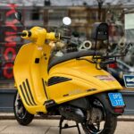 Gele AGM-scooter met blauwe kentekenplaat en rugsteun, geparkeerd voor een winkelruit.