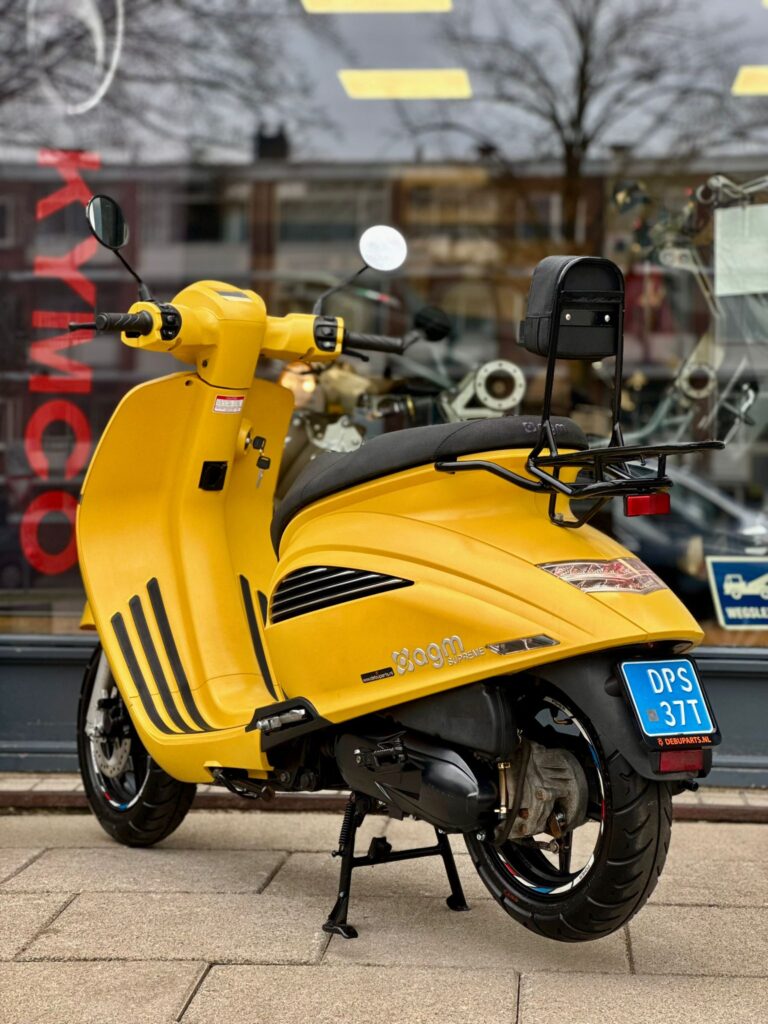 Gele AGM-scooter met blauwe kentekenplaat en rugsteun, geparkeerd voor een winkelruit.